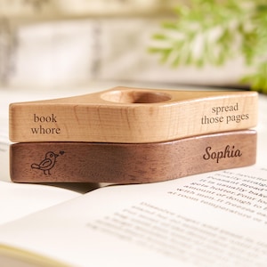 Puede incluir: Soporte de libro de madera con las inscripciones "book whore" y "spread those pages" grabadas en la parte superior. La pieza inferior est&aacute; grabada con el nombre "Sophia" y una ilustraci&oacute;n de un p&aacute;jaro. El soporte est&aacute; sobre un libro abierto.