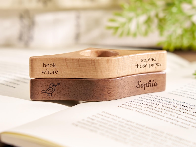 Puede incluir: Soporte de libro de madera con las inscripciones "book whore" y "spread those pages" grabadas en la parte superior. La pieza inferior est&aacute; grabada con el nombre "Sophia" y una ilustraci&oacute;n de un p&aacute;jaro. El soporte est&aacute; sobre un libro abierto.