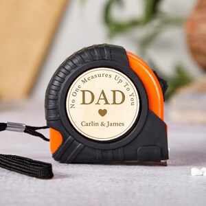 May include: An orange and black tape measure with the text "DAD" and "No One Measures Up To You" on a wooden face. The tape measure has a black wrist strap and the names "Carlin & James" are also visible.