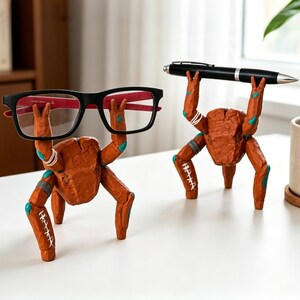 May include: Two orange, textured, creature-shaped desk organizers with multiple limbs. One figure holds black-framed eyeglasses with red inner temples, while the other figure holds a black ballpoint pen horizontally above its head.