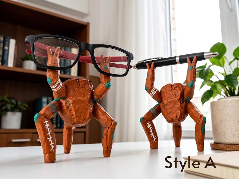May include: Two orange, textured, multi-legged creature figurines acting as holders. One holds black-framed glasses with red inner temples, and the other holds a black ballpoint pen. Each figure features white stitched-like markings and teal accents. Style A.