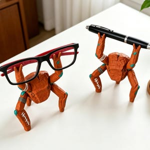 May include: Two orange, textured, crab-shaped desk organizers with articulated limbs. One crab holds a pair of black and red eyeglasses, while the other crab holds a black ballpoint pen. Both figures feature white stitched details on their legs.