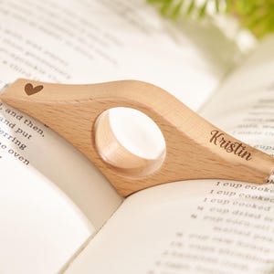 Puede incluir: Un sujetap&aacute;ginas de madera clara con un coraz&oacute;n grabado y el nombre "Kristin". Tiene una abertura circular y est&aacute; colocado sobre las p&aacute;ginas abiertas de un libro.