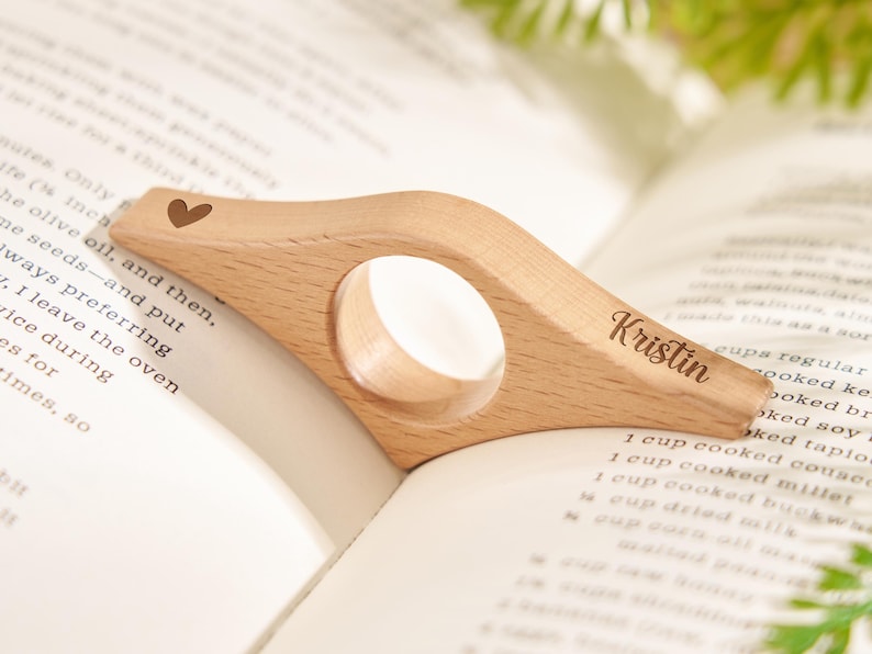 Puede incluir: Un sujetap&aacute;ginas de madera clara con un coraz&oacute;n grabado y el nombre "Kristin". Tiene una abertura circular y est&aacute; colocado sobre las p&aacute;ginas abiertas de un libro.