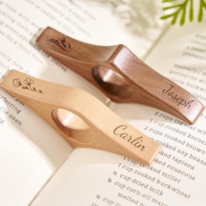 Puede incluir: Dos sujetap&aacute;ginas de madera, uno claro y otro oscuro, descansan sobre un libro. El soporte de color claro tiene grabado el nombre "Carlin" y un dise&ntilde;o floral. El soporte oscuro tiene el nombre "Joseph" y un dise&ntilde;o de monta&ntilde;a.