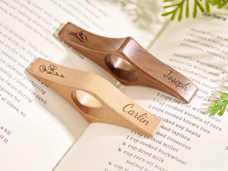 Puede incluir: Dos sujetap&aacute;ginas de madera, uno claro y otro oscuro, descansan sobre un libro. El soporte de color claro tiene grabado el nombre "Carlin" y un dise&ntilde;o floral. El soporte oscuro tiene el nombre "Joseph" y un dise&ntilde;o de monta&ntilde;a.
