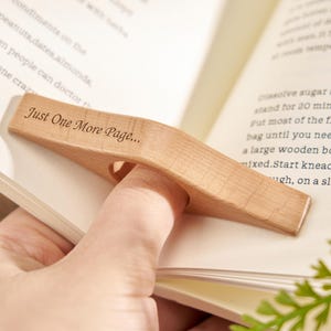 Puede incluir: Un sujetap&aacute;ginas de madera clara con la inscripci&oacute;n "Just One More Page...". El sujetap&aacute;ginas est&aacute; dise&ntilde;ado para colocarse sobre un dedo, manteniendo un libro abierto. El libro est&aacute; abierto en una p&aacute;gina con texto.