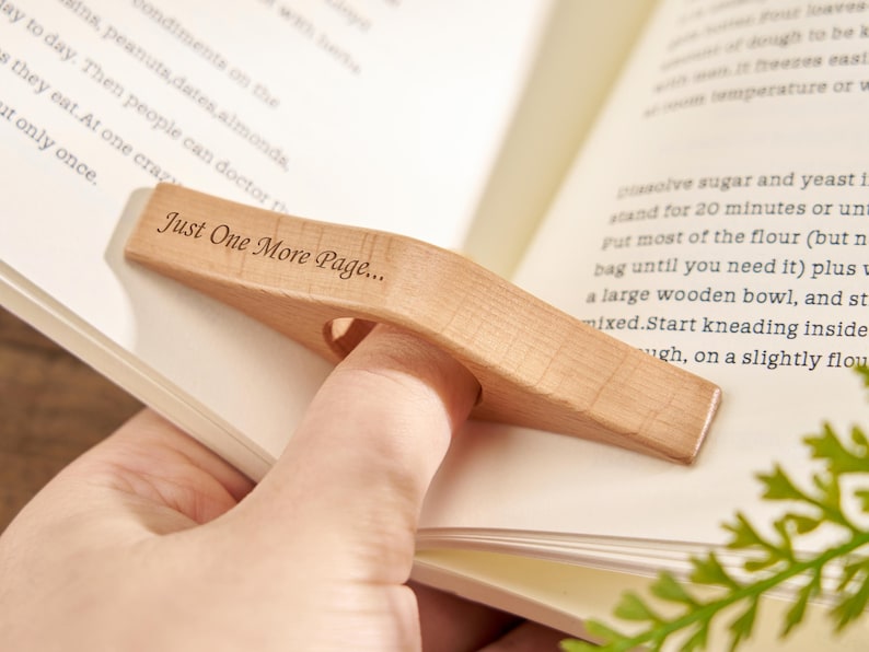 Puede incluir: Un sujetap&aacute;ginas de madera clara con la inscripci&oacute;n "Just One More Page...". El sujetap&aacute;ginas est&aacute; dise&ntilde;ado para colocarse sobre un dedo, manteniendo un libro abierto. El libro est&aacute; abierto en una p&aacute;gina con texto.