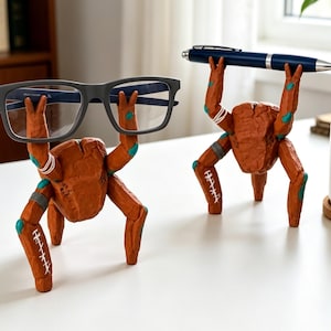 May include: Two orange, textured, multi-legged creature figurines acting as holders. One figurine supports a pair of dark gray eyeglasses, while the other holds a dark blue ballpoint pen. Both figures feature teal accents and white stitched details.