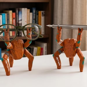 May include: Two brown, carved-style figures with textured surfaces and teal accents, each standing on four legs. One figure holds a pair of olive green glasses, while the other holds a silver fountain pen horizontally above its head.