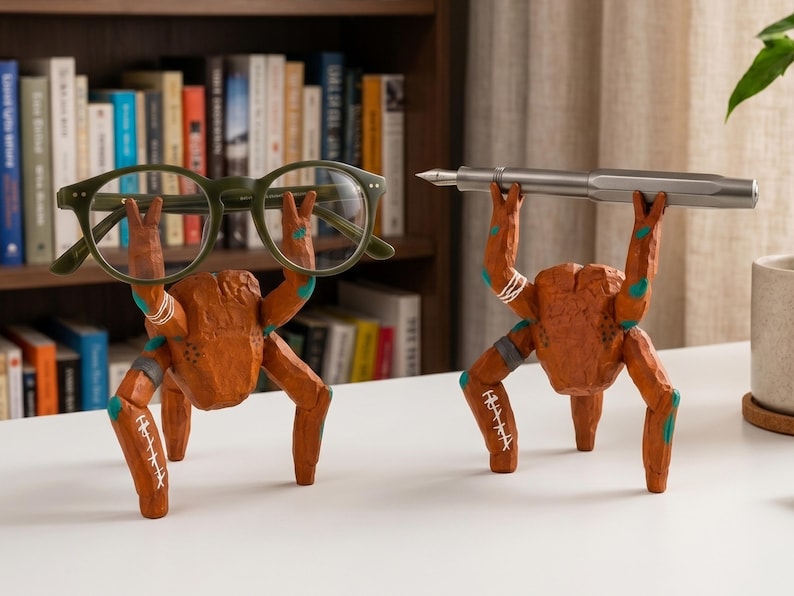 May include: Two brown, carved-style crab figurines with textured surfaces and teal accents. One crab holds a pair of olive green eyeglasses with its raised claws, while the other crab holds a silver fountain pen horizontally across its raised claws.