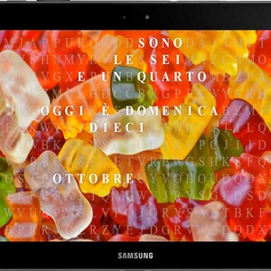 Könnte beinhalten: Ein Samsung-Tablet zeigt ein farbenfrohes Bild von Gummibärchen mit überlagertem Text. Der Text, auf Italienisch, lautet: "Sono le sei e un quarto. Oggi è domenica dieci ottobre."