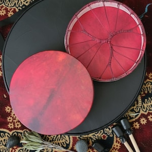 May include: Two red hand drums with a black case and drumsticks. One drum is plain, the other has a tree-like design. The drums are on a patterned rug with stones and greenery.