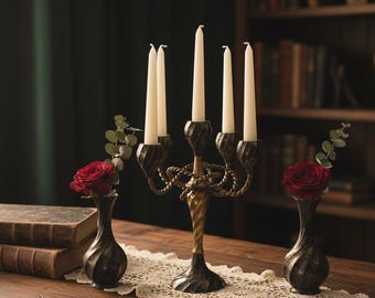Puede incluir: Un candelabro decorativo con cinco velas blancas, flanqueado por dos pequeños jarrones con rosas rojas. El candelabro tiene un acabado de bronce con un tallo retorcido y detalles de cuerda. También se ve un camino de mesa de encaje y libros.