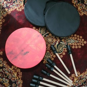 Puede incluir: Un conjunto de instrumentos de percusi&oacute;n, que incluye un tambor rojo, fundas negras para tambor y baquetas. El tambor es redondo con una superficie de color rosa rojizo. Las baquetas tienen mangos de madera y extremos de golpeo acolchados negros. Las fundas son negras y circulares.
