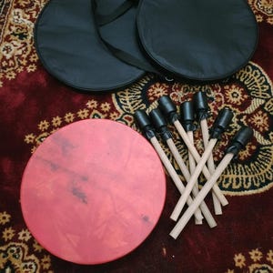 Puede incluir: Un tambor rojo de forma circular, acompa&ntilde;ado de un juego de baquetas y dos fundas de transporte negras. Las baquetas tienen mangos de madera y puntas negras. Los elementos est&aacute;n colocados sobre una alfombra marr&oacute;n rojiza estampada.