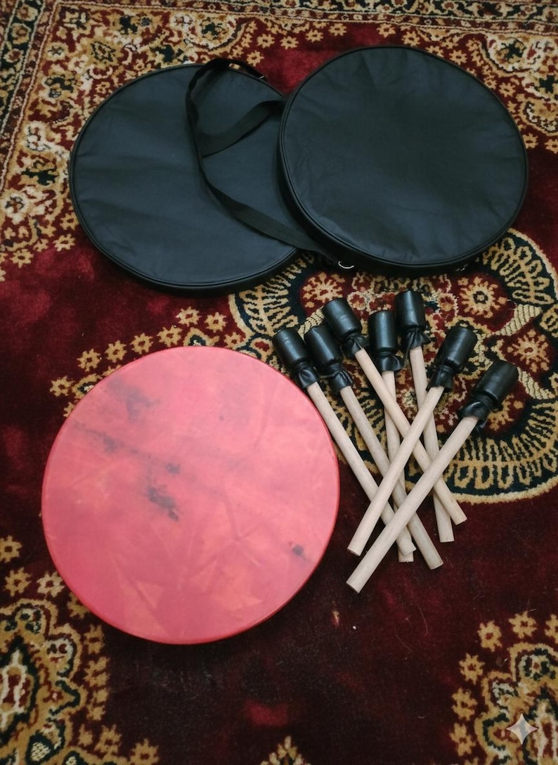 Puede incluir: Un tambor rojo de forma circular, acompa&ntilde;ado de un juego de baquetas y dos fundas de transporte negras. Las baquetas tienen mangos de madera y puntas negras. Los elementos est&aacute;n colocados sobre una alfombra marr&oacute;n rojiza estampada.