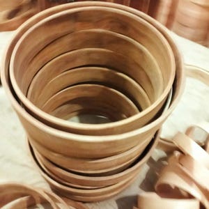 May include: A stack of unfinished wooden bowls in various sizes. The bowls are made of light brown wood and have a smooth, natural finish. The bowls are nested together, with the largest bowl at the bottom and the smallest at the top.