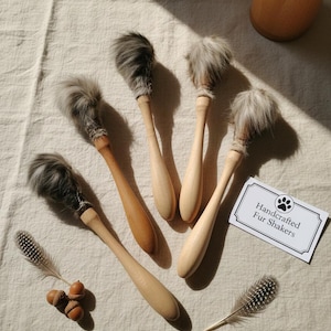 Op de afbeelding: Een verzameling handgemaakte fur shakers met houten handvatten en pluizige grijze vachttoppen. De shakers liggen op een crèmekleurige doek, met enkele veren en eikels eromheen. Een kaartje leest "Handcrafted Fur Shakers".