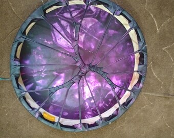 Puede incluir: Un objeto circular morado y negro con un diseño único. Tiene un borde oscuro y un área central con un tono morado vibrante. Finas líneas oscuras irradian desde el centro, creando un patrón segmentado. El objeto parece estar hecho de tela.