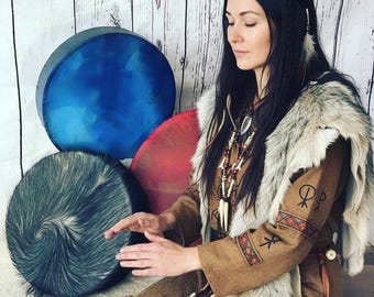 Puede incluir: Una persona con un vestido marrón y un chal de piel se sienta cerca de tres tambores. Los tambores son azules, rojos y grises. Una maza de tambor de madera descansa en el suelo. La persona lleva un collar con cuentas y colgantes.