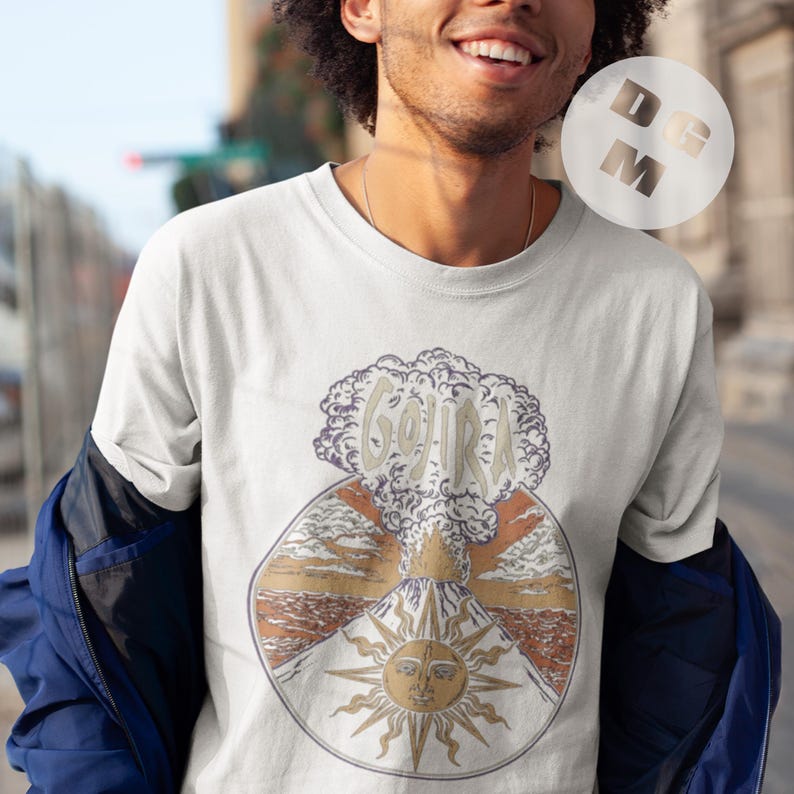 May include: White t-shirt with a graphic print of a volcano erupting with the word "GOJIRA" in the smoke. The volcano is surrounded by a sun with a face.