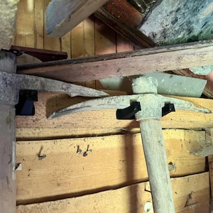 May include: Two weathered pickaxes with wooden handles and metal heads are mounted horizontally on a wooden wall using black plastic brackets. The metal heads show signs of age and oxidation, while the wooden handles appear worn and textured.