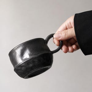 Può includere: Una tazza in ceramica nera con una superficie testurizzata e irregolare, caratterizzata da una finitura liscia e lucida sul bordo superiore. La tazza presenta un manico arrotondato e una forma artigianale, con un colore grigio antracite.