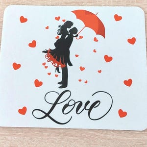 Könnte beinhalten: Weißes rechteckiges Mousepad mit einer schwarzen Silhouette eines Paares, das sich unter einem roten Regenschirm umarmt, umgeben von roten Herzen. Das Wort "Love" ist in eleganter Schreibschrift unten geschrieben. Romantisches Thema.