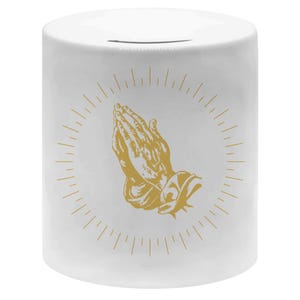 Könnte beinhalten: Weiße Keramik-Spardose mit einer goldenen Illustration von betenden Händen, umgeben von strahlenförmigen Linien. Die Spardose hat oben einen Schlitz zum Einwerfen von Geld.