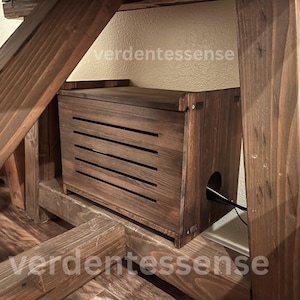 May include: A dark stained wooden box with horizontal slats on the front, positioned under a wooden structure. The box has a lid and a hole for a cable. The overall design suggests a storage solution or a decorative element.