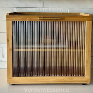 May include: A rectangular bamboo bread box with a clear, ribbed glass door and a brass-colored handle. The box has a flat top and is sitting on a white countertop. The brand name "VerdentEssence" is visible in the image.