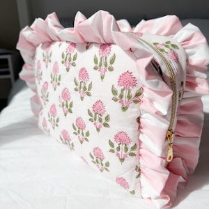 May include: A rectangular quilted pouch featuring a white fabric base with a repeating pink floral and green leaf block print pattern. The pouch is bordered by a wide, ruffled trim in a pink and white striped fabric and includes a gold zipper.