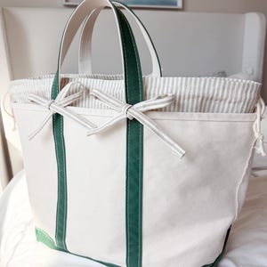 May include: A cream-colored canvas tote bag with green accents. The bag features green handles and trim, with a striped fabric lining and drawstring closure. Two decorative bows are tied on the front. The bag is sitting on a white surface.