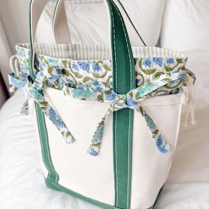May include: A cream-coloured canvas tote bag featuring dark green vertical trim and handles. The bag includes a drawstring closure made from a white and blue floral patterned fabric, which is tied into bows on the front exterior of the bag.