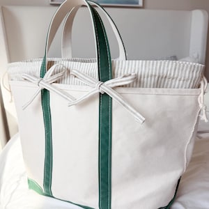 May include: A cream-coloured canvas tote bag featuring dark green vertical stripes and a matching green base. The bag includes a beige and white striped drawstring closure at the top, accented with two decorative bows tied at the front.