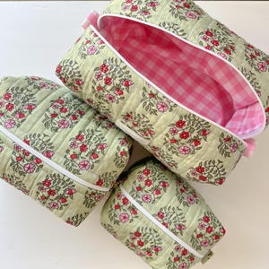 May include: Three quilted pouches in varying sizes, featuring a light green fabric with a floral pattern of red and pink blossoms and green leaves. Each pouch has a white zipper closure and a pink and white gingham fabric lining inside.