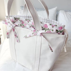 May include: A cream-colored canvas tote bag featuring two sturdy handles and a drawstring closure. The interior lining displays a white fabric with a pink floral pattern and green stems, secured by two fabric ties on the sides of the bag.