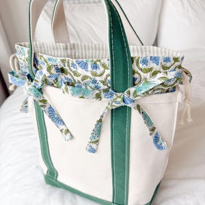 May include: A white canvas tote bag with green trim, base, and handles. The interior lining is a floral fabric with blue and green flowers. The top edge is gathered and tied with floral fabric bows. The bag is designed for carrying items.