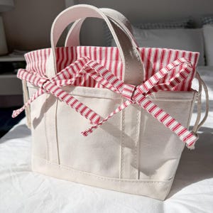 May include: A small off-white canvas tote bag featuring two sturdy handles and a red and white striped fabric insert. The insert extends from the top and is cinched with matching red and white striped fabric ties that are knotted into a bow.