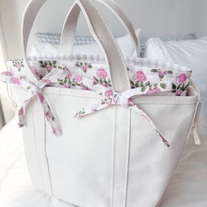 May include: A white canvas tote bag with sturdy handles. The interior features a floral print with pink flowers and green leaves. The top edge is trimmed with a gingham pattern. Decorative bows with the floral print accent the exterior.