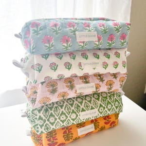 Peut inclure: Une pile de cinq trousses à crayons rectangulaires en tissu matelassé avec fermetures éclair. Chaque trousse présente un imprimé floral ou géométrique unique en bleu, blanc, rose, vert ou orange, et une étiquette blanche avec LITTLE GOOSE.