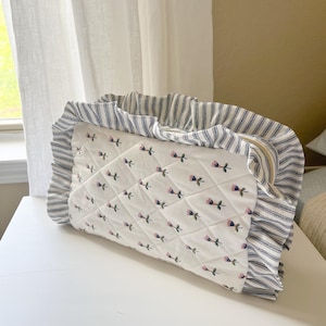 A white and blue quilted bag with a floral pattern sits on a white surface, with a window and curtain visible in the background.