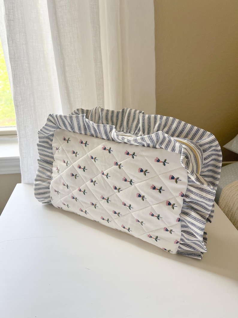A white and blue quilted bag with a floral pattern sits on a white surface, with a window and curtain visible in the background.
