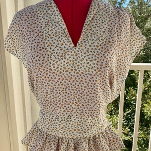 Puede incluir: Una blusa blanca con cuello en V y mangas cortas, adornada con un delicado estampado floral en amarillo y verde. La blusa presenta una cintura fruncida y un dobladillo con volantes, exhibida en un maniquí rojo.