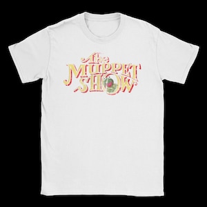 Peut inclure: Un t-shirt blanc à manches courtes avec une impression graphique vieillie sur la poitrine. Le motif affiche le texte The Muppet Show dans une police stylisée jaune et rouge, avec une petite image circulaire de Kermit la grenouille dans le O.
