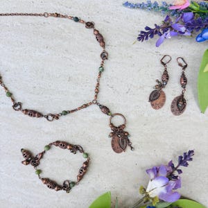 Puede incluir: Un conjunto de joyas en tono cobre que incluye collar, pulsera y pendientes. El collar y la pulsera incorporan cuentas verdes. Los pendientes tienen colgantes ovalados con diseños de hojas y relojes. El conjunto se exhibe sobre una superficie clara con detalles florales.