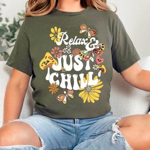 Puede incluir: Una camiseta verde con un diseño gráfico retro que presenta el texto "Relax & Just Chill" rodeado de flores y mariposas.