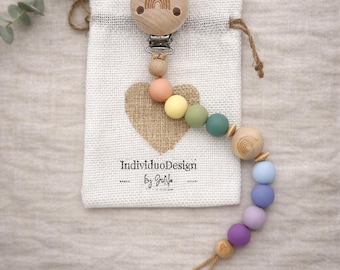 May include: A wooden pacifier clip with a rainbow design and a string of colorful beads. The clip is attached to a small, off-white drawstring bag with a heart design and the text "IndividuoDesign by GioLife".