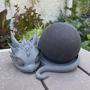 以下が含まれることがあります： スパイクのある詳細な頭部と曲がった尾を持つ、グレーの3Dプリント製ドラゴン型スタンド。球体のチャコールグレーのスマートスピーカーを保持するように設計されています。ドラゴンのフィギュアはスピーカーの横に配置されています。